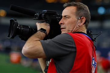 A man with a video camera is wearing a red vest with the NFL logo on the back.