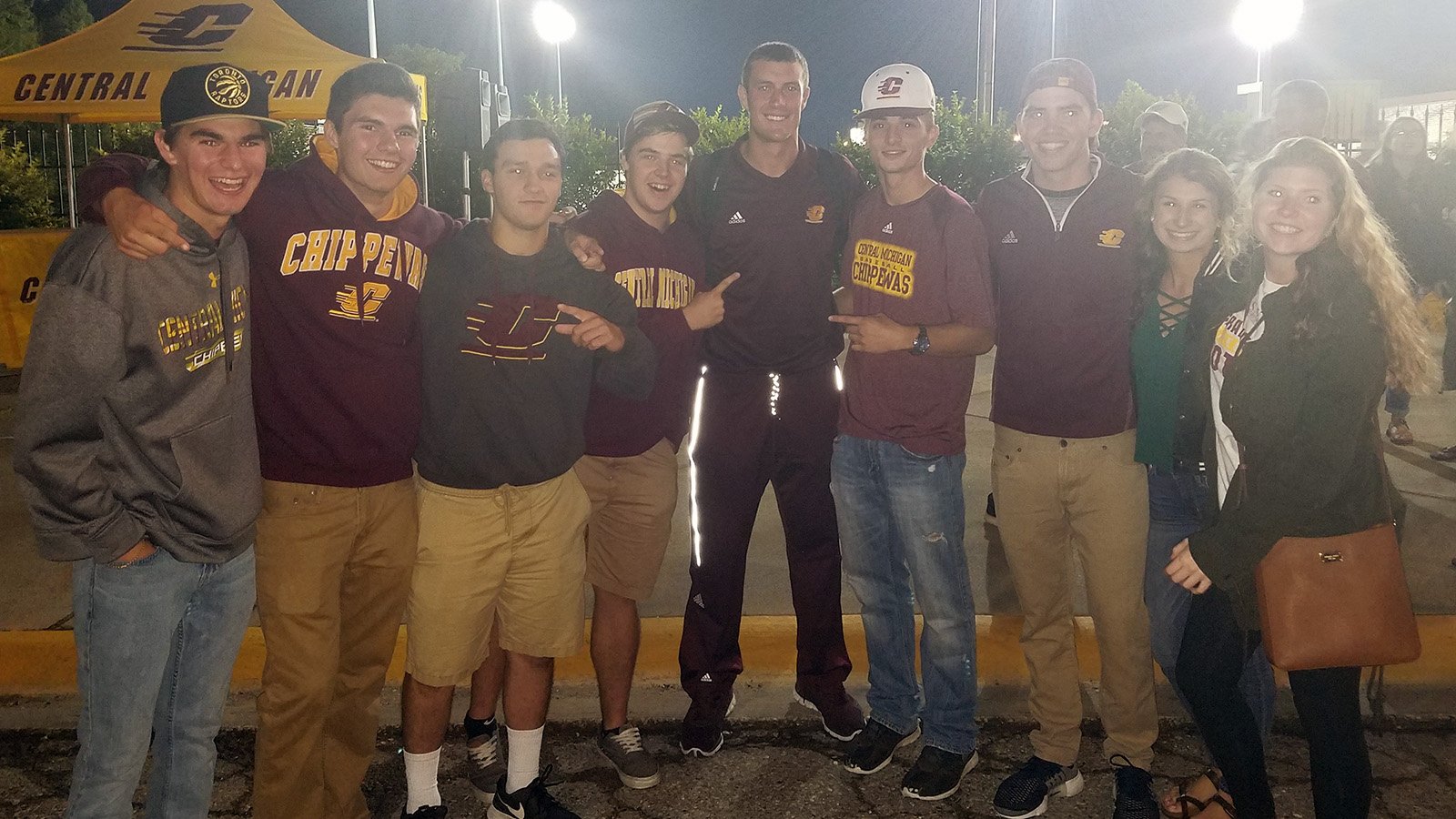 A group of eight CMU students wearing CMU gear pose with football player Jesse Kroll in the middle outside of a football stadium at night.