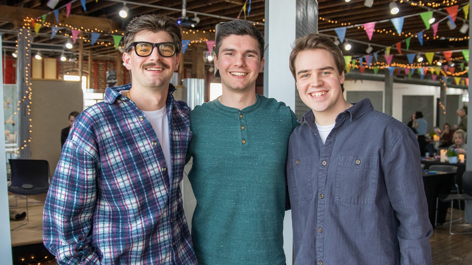 Three young men pose for a picture. Each has brown hair. The one on the left is wearing glasses and a plaid shirt. The one in the middle wears a green henley shirt, and the one on the right wears a navy blue long-sleeve shirt.