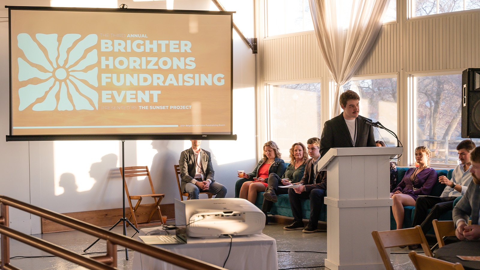 A young man with brown hair and a mustache speaks behind a white podium with a group of people behind him on a couch. A sign over his shoulder reads Brighter Horizons Fundraising Event.