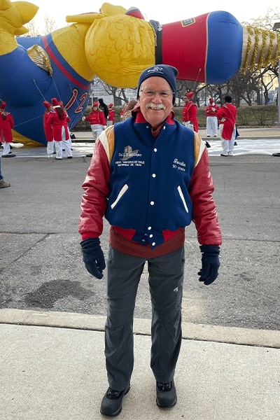 A man wearing a blue letterman's jacket, blue winter hat, and winter gloves poses in front of the Big Bird balloon at America's Thanksgiving Parade.