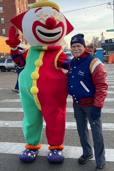 A man wearing a blue letterman's jacket, blue winter hat, and winter gloves poses with Clownie at America's Thanksgiving Parade.