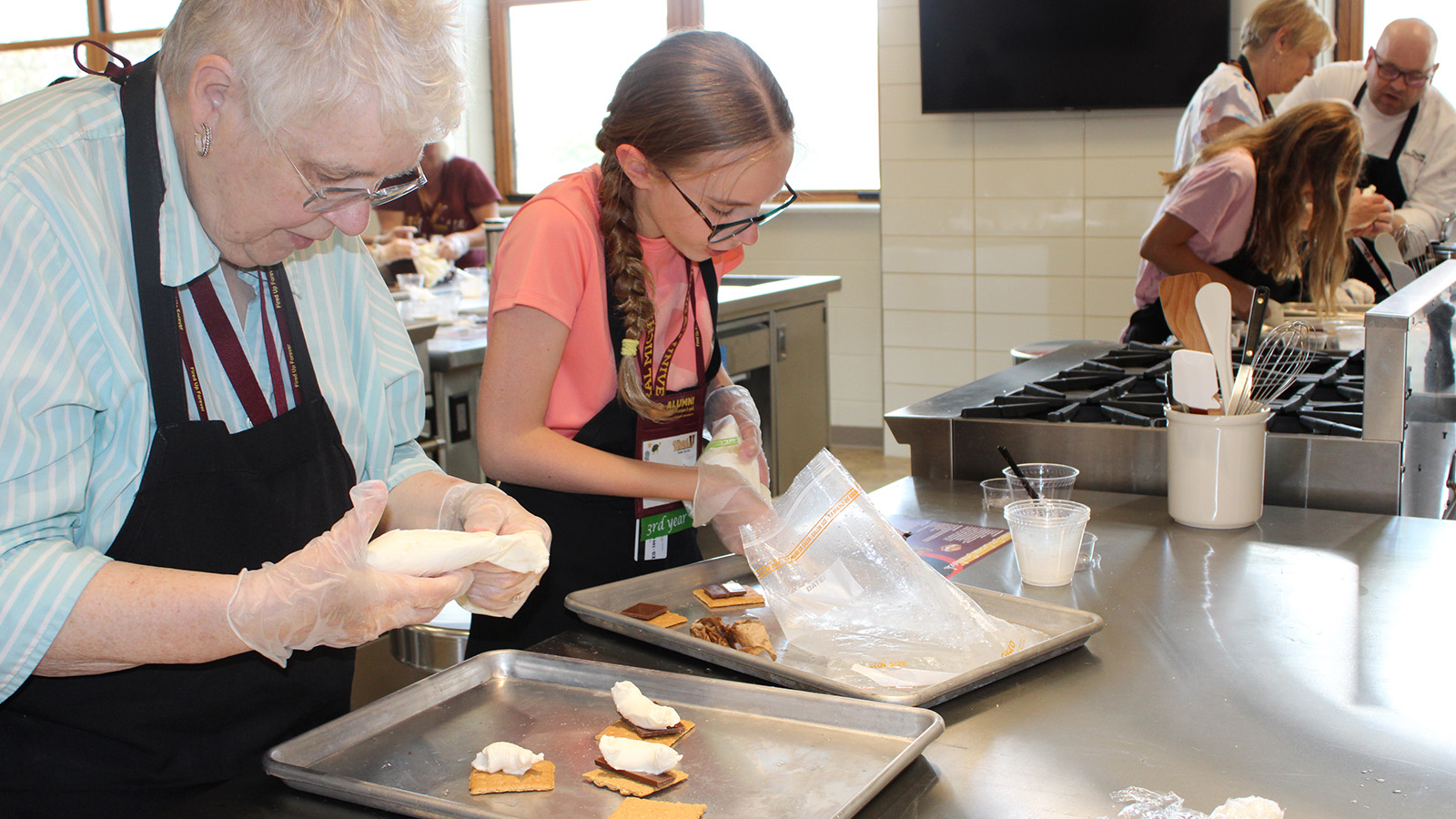 In the foreground, a grandmother and her granddaughter are wearing aprons in a kitchen and making smores. In the background, a chef helps another grandmother and her granddaughter with their smores.