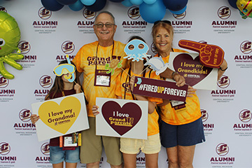 Two grandparents and their two grandchildren, wearing gold and maroon t-shirts, pose in front of an alumni association backdrop. They hold up signs that say I love my Grandma, I love Grandparents U, #FiredUpForever, and I love my Grandkids.