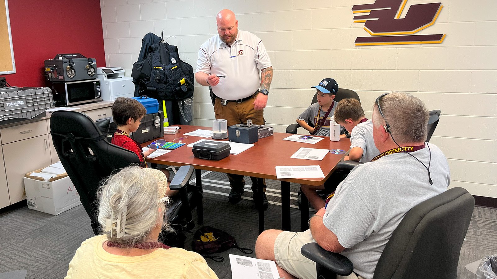 Two grandparents sit in the foreground while their three grandchildren sit around a table in the background, watching a police officer dressed in a CMU polo shirt teach them how to take fingerprints.