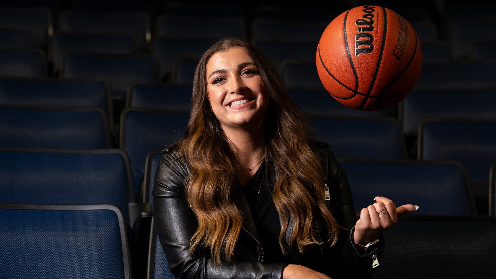 A woman in a black leather jacket smiles and tosses a basketball in the air while sitting.