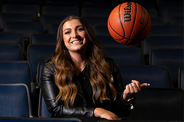 A woman in a black leather jacket smiles and tosses a basketball in the air while sitting.