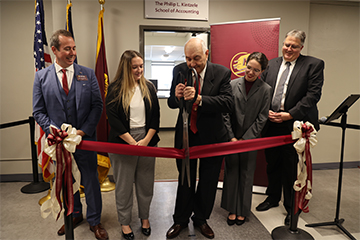 A man cutting a red ribbon with a pair of scissors while two men and two women stand next to him and watch.