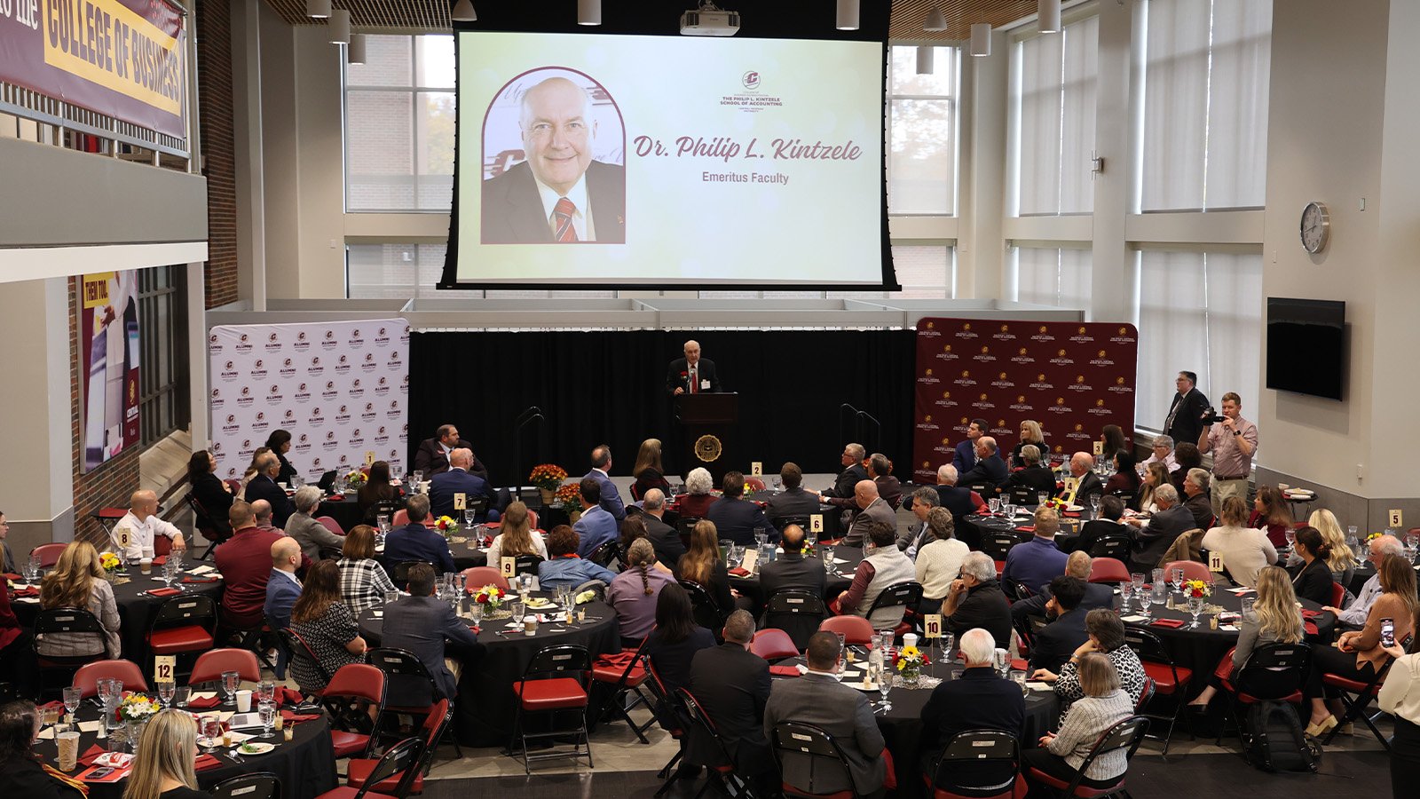 A group of people sit at tables during a luncheon while a man stands on a stage while speaking in front of a podium.