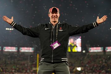 A man wearing a black zipped-up jacket and red hat stands and smiles with his arms outstretched.