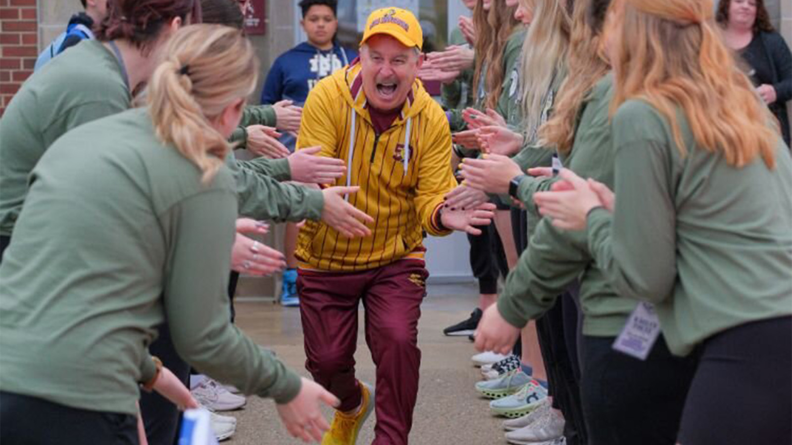 A man wearing a maroon and gold jumpsuit and ballcap happily gives high-fives while running through a human-formed tunnel of college students dressed in light green shirts.