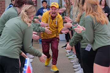 A man wearing a maroon and gold jumpsuit and ball cap happily gives high-fives while running through a human-formed tunnel of college students dressed in light green shirts.