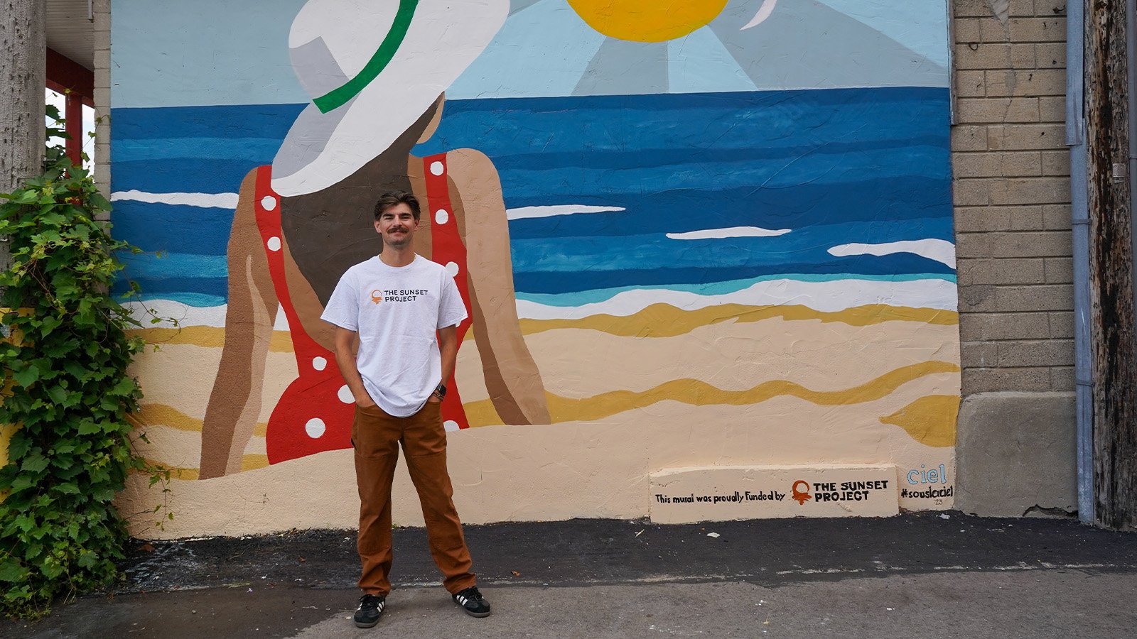 A young man with brown hair and a mustache, wearing dark brown khakis and a white t-shirt, stands in front of a painted mural of a woman at the beach looking at the sun.