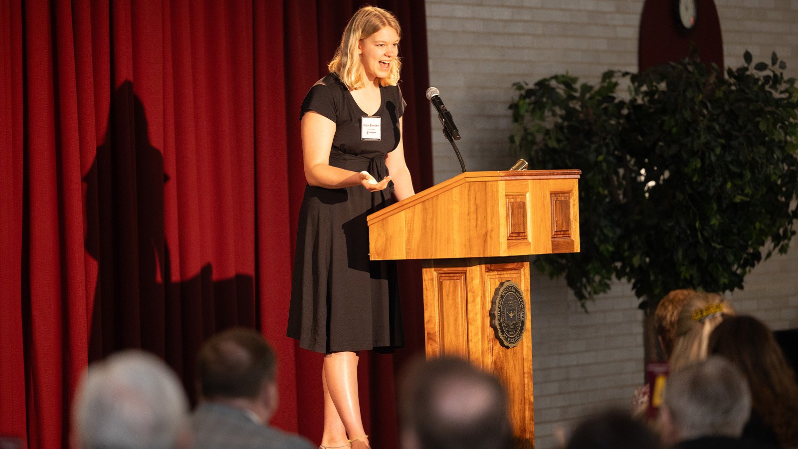 A young woman with light brown hair wearing a black dress is on a stage standing behind a podium and speaking in front of a crowded room.