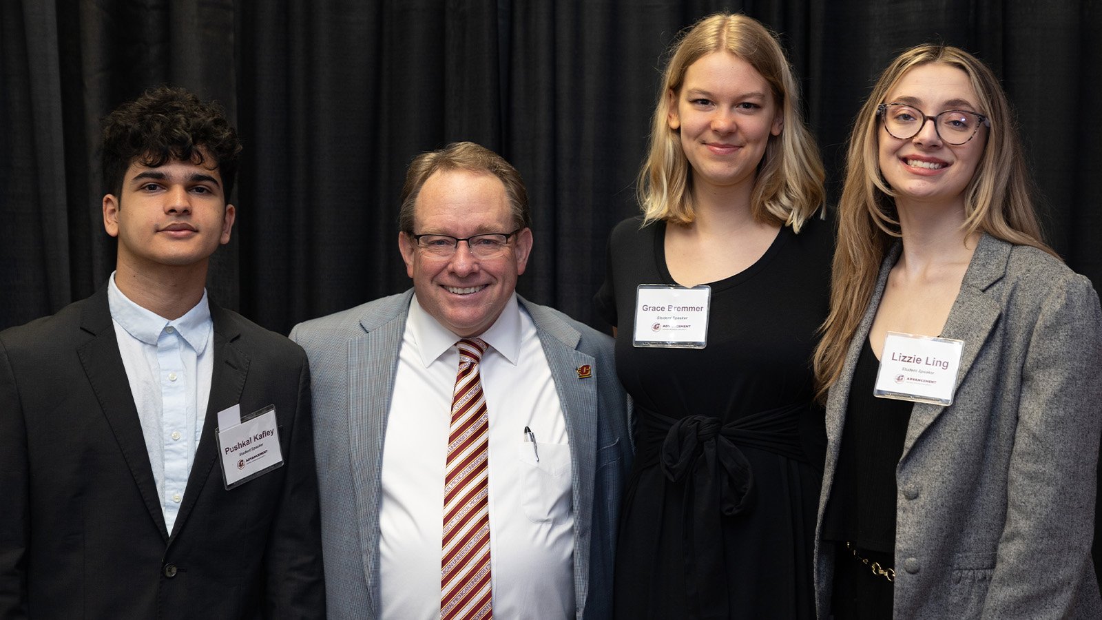 A man with glasses wearing a grey suit, white shirt, and striped tie stands among three college students—one male with dark hair and a dark suit and two females with light brown hair. One wears a black dress the other has a grey jacket over a dark shirt.