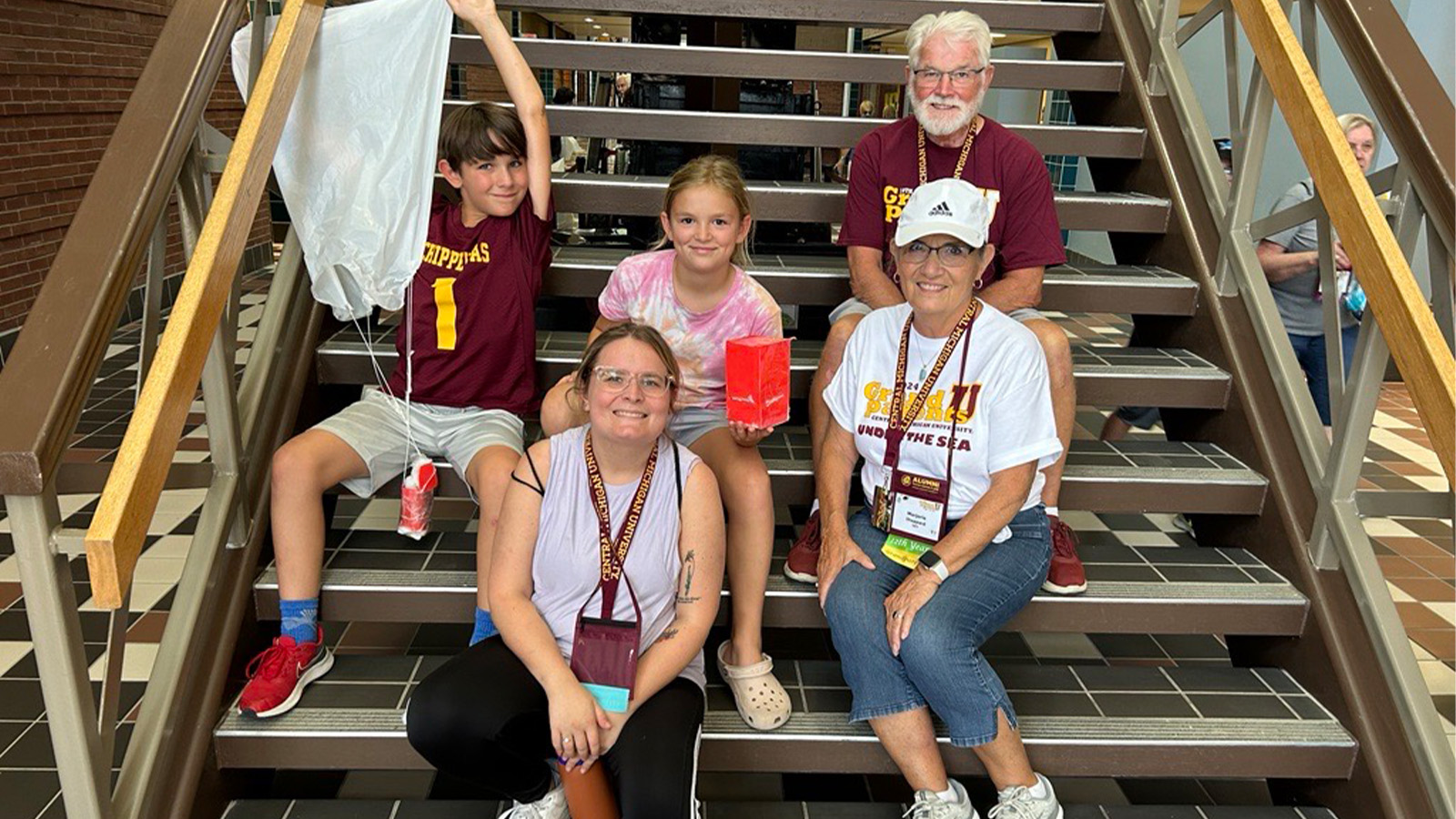 A family of five, including two grandparents and three grandchildren, sit on a staircase and smile. The boy grandchild is holding a parachute with a small red box attached to it. The girl grandchild holds a small red box.