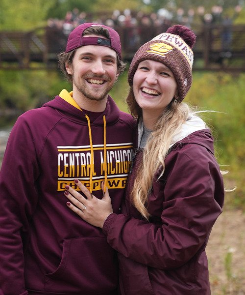 A man wearing a CMU hooded sweatshirt and backward ball cap smiles and poses with a woman wearing a maroon jacket and CMU winter hat while showing off her engagement ring.