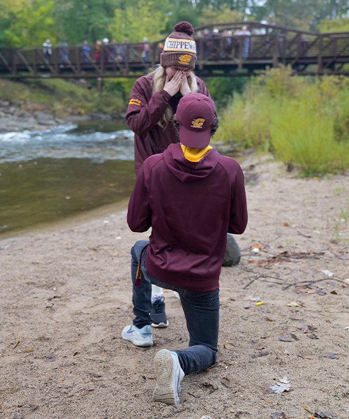 A man wearing a maroon hooded sweatshirt and backward action C ball cap is down on one knee in the sand beside a river, proposing to a woman wearing a maroon jacket and CMU winter hat.