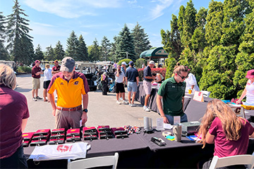 In the foreground, two men in golf attire are looking at items on a table on the pavement near the clubhouse of a golf course while groups of golfers converse in the background.