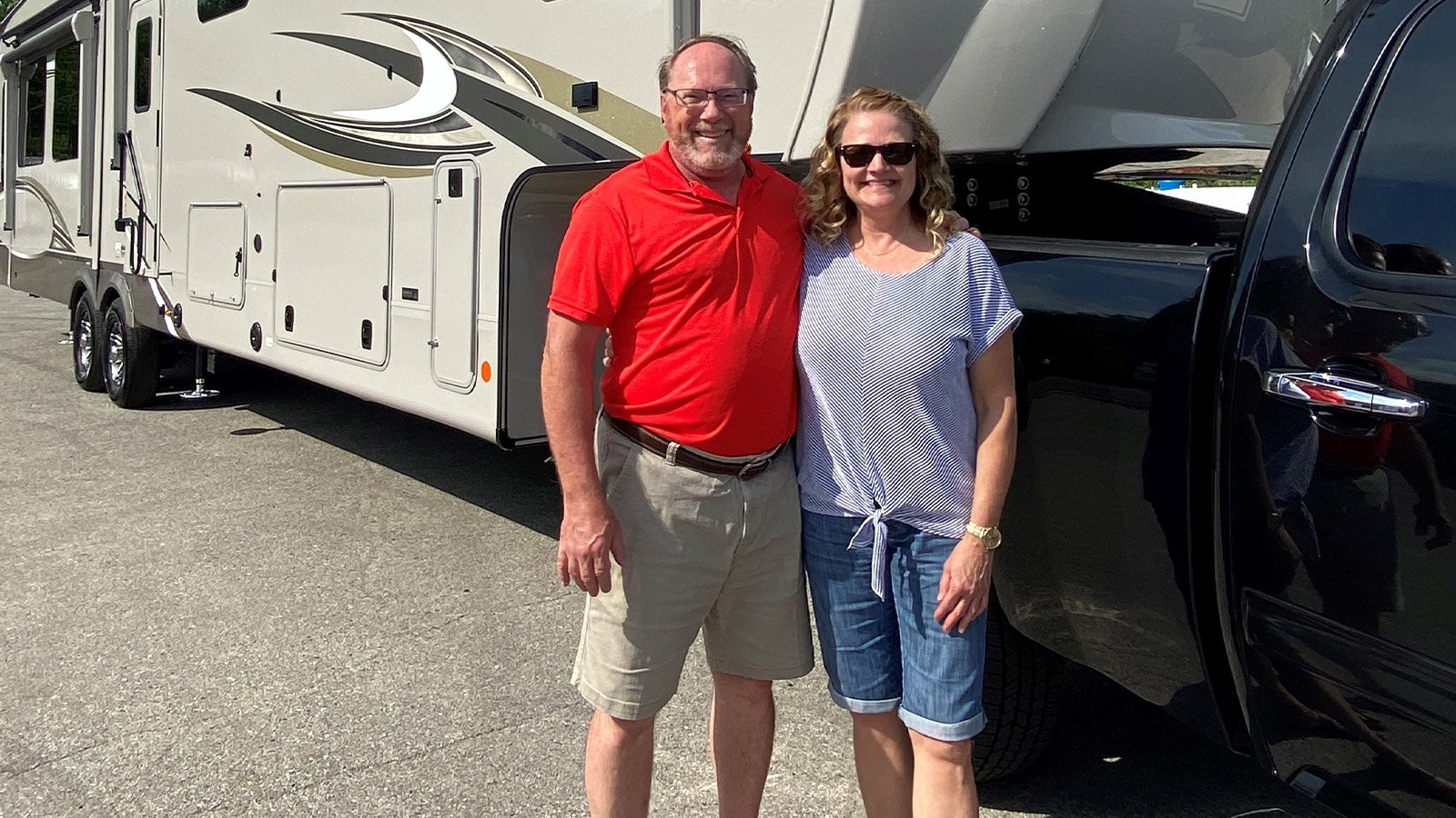 A man in a red polo and a woman in summer clothing stand in front of a RV