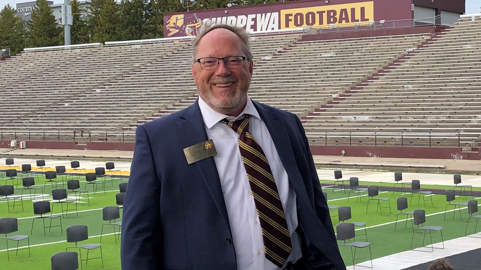 A man with glasses wears a white dress shirt and striped tie under a blue sports jacket while standing in front of chairs on the Central Michigan University football field.