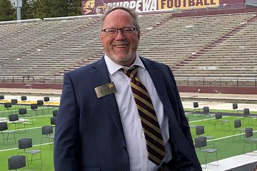 A man with glasses wears a white dress shirt and striped tie under a blue sports jacket while standing in front of chairs on the Central Michigan University football field.