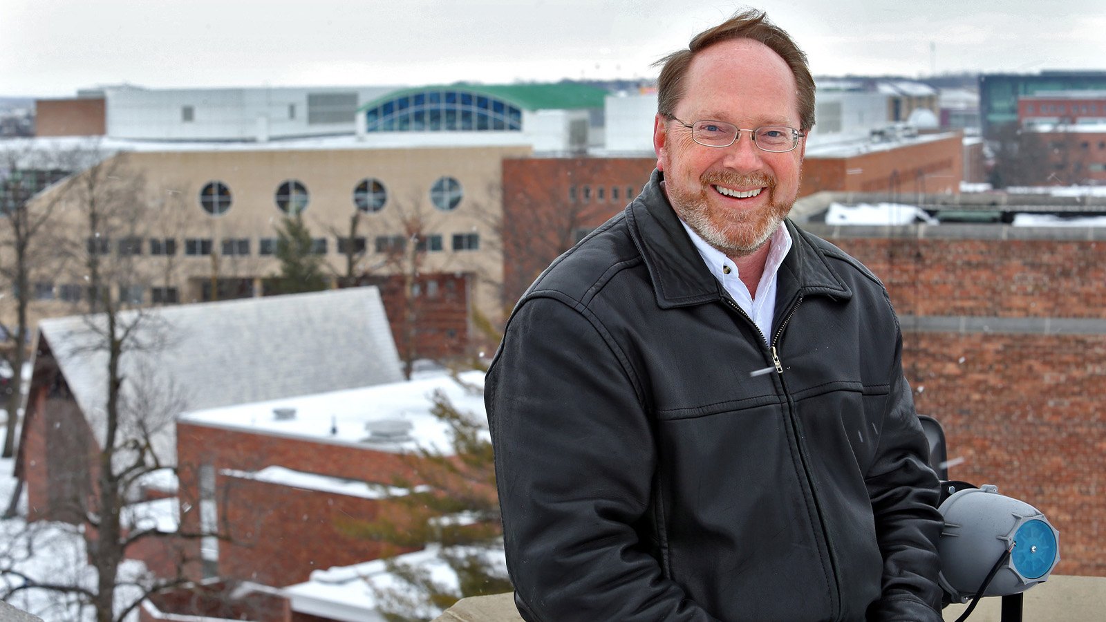 A man in a black leather jacket poses while sitting on a rooftop with buildings in the distance behind him.